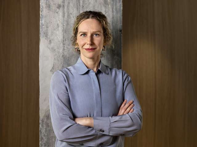 Sabine Müller steht in hellblauem Hemd vor einer Wand.